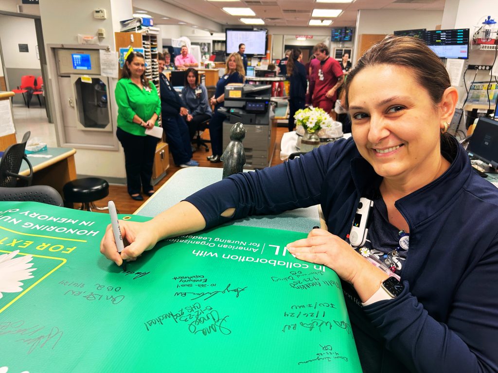 A woman smiles while signing a large green banner at a busy hospital station, surrounded by staff members in the background.