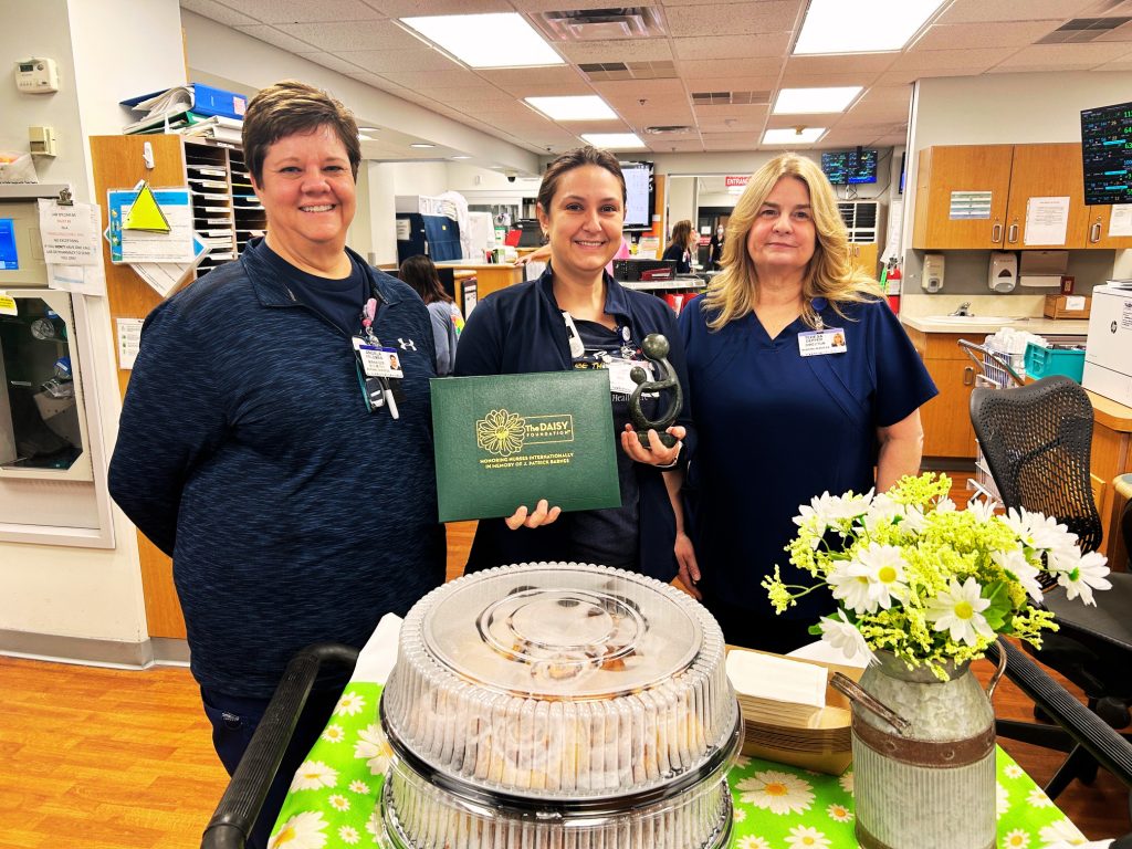 Three smiling healthcare workers stand behind a table with a cake, flowers, and an award in a hospital setting. One person holds a green certificate and a glass trophy.