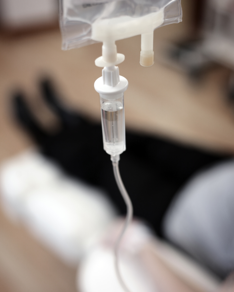 Close-up of an IV drip with clear fluid, with a person lying on a bed in the blurred background, suggesting a medical or hospital setting.