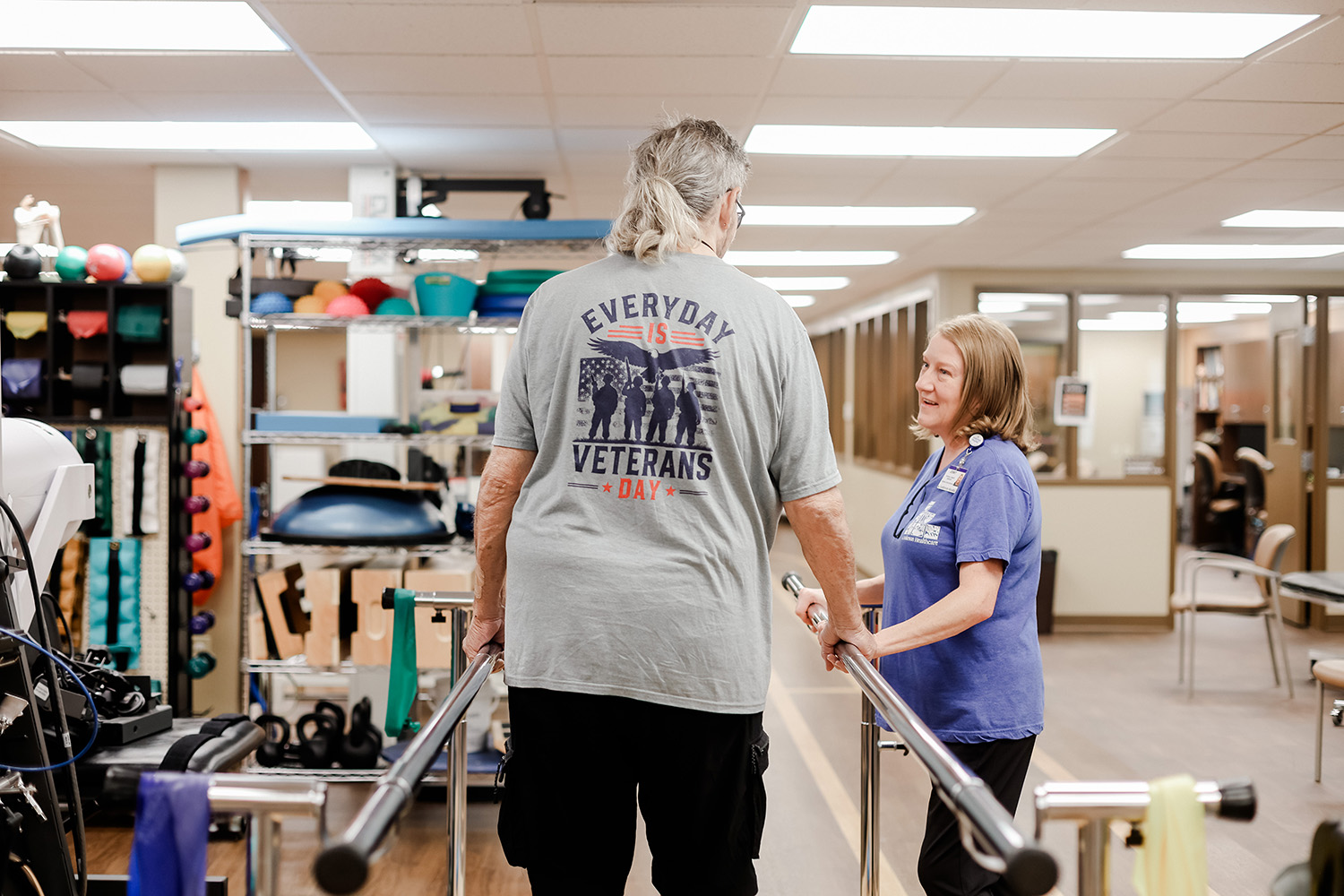 An older man walking between parallel bars in a therapy gym, assisted by a smiling female therapist. The man wears a shirt with Every Day is Veterans Day printed on the back.